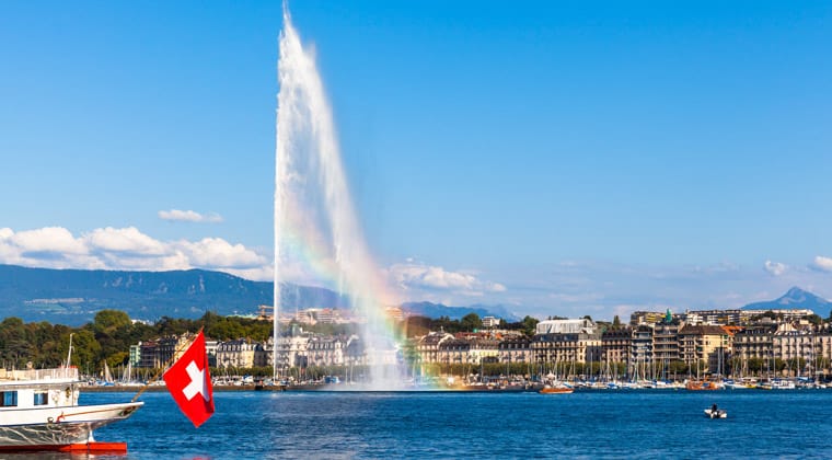 Blick vom Wasser aus auf die Stadt Genf mit Wasserfontäne und Alpenpanorama