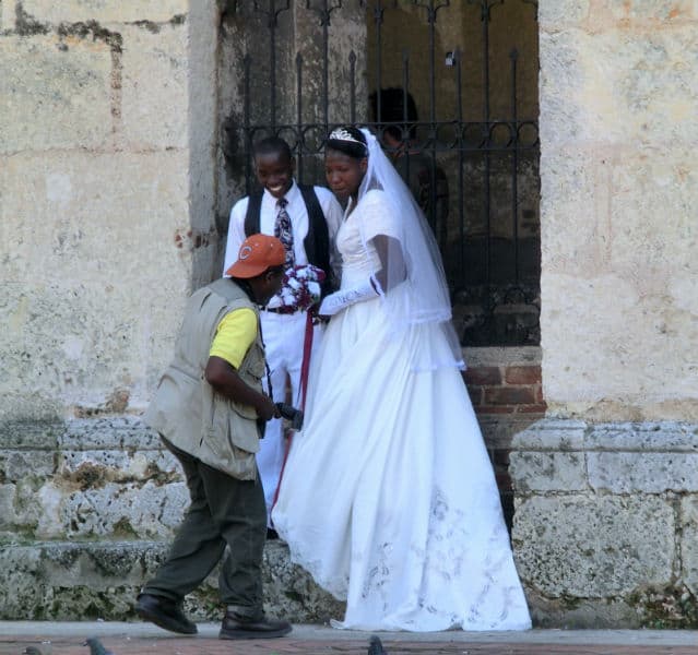 Dominikanisches Hochzeitspaar vor einer rustikalen Kirchenmauer mit einem Fotografen um Vordergrund