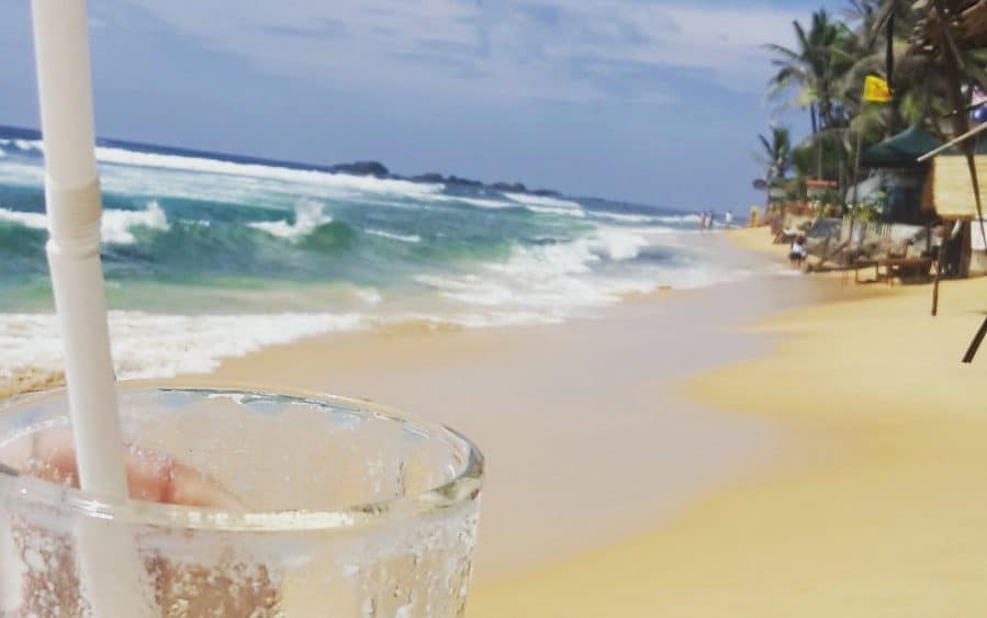 Strand Arugam Bay in Sri Lanka, heller Sandstrand, türkisfarbenes Meer, Wellen mit weißer Gischt, grüne Palme, Strandbar, helles Glas mit Strohhalm