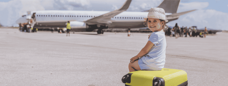 Kleines lächelndes Mädchen in Sommerkleidung, das auf einem gelben Koffer auf einem Rollfeld am Flughafen sitzt, im Hintergrund ein Flugzeug