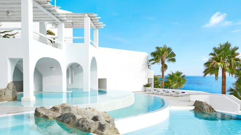 Weißes Hotel mit hellblauem Pool, Palmen und Terrasse mit Liegestühlen direkt am blauen Meer.