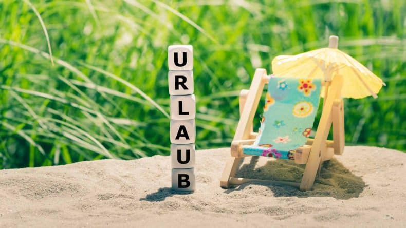 Am Strand bilden Würfeln das Wort "Urlaub" neben einer Sonnenliege und einem Sonnenschirm.