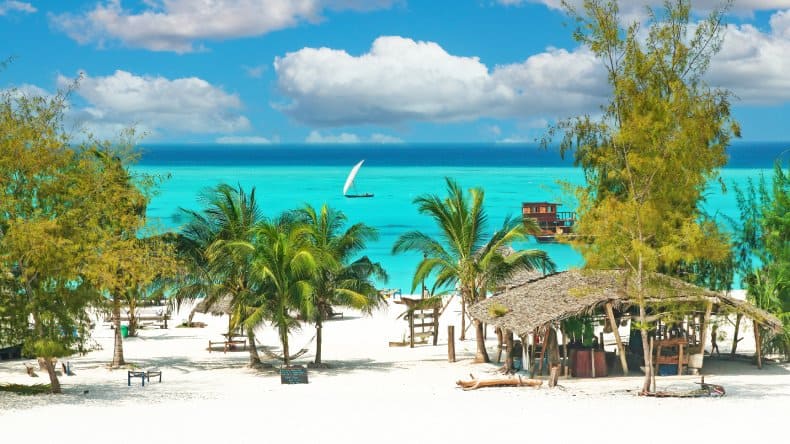 Blick auf einen weißen Sandstrand mit Palmen und grümnen Bäumen, Hängematten, einer Strandbar und türkisfarbenes Meer, Insel Sansibar, Indischer Ozean.