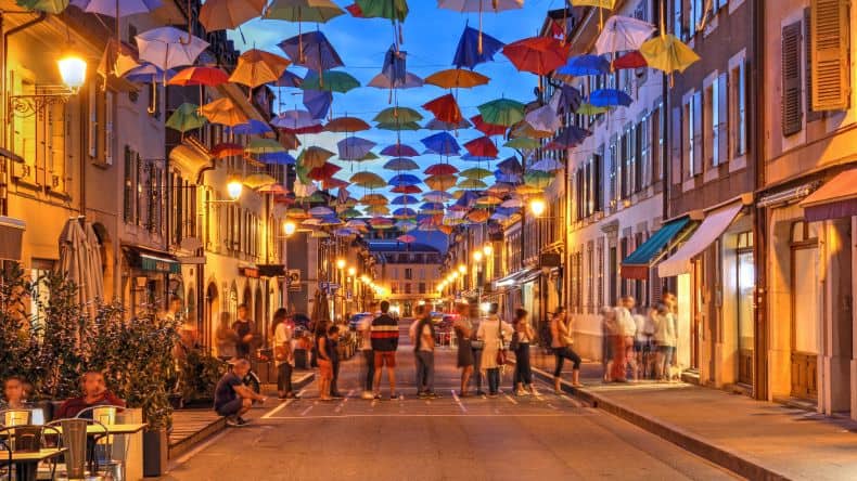 Blick in eine Straße im Künstlerort Courage am Abend, Straßenbeleuchtung, Menschen, bunte Regenschirme über der Strasse, Genf, Schweiz, Frankreich