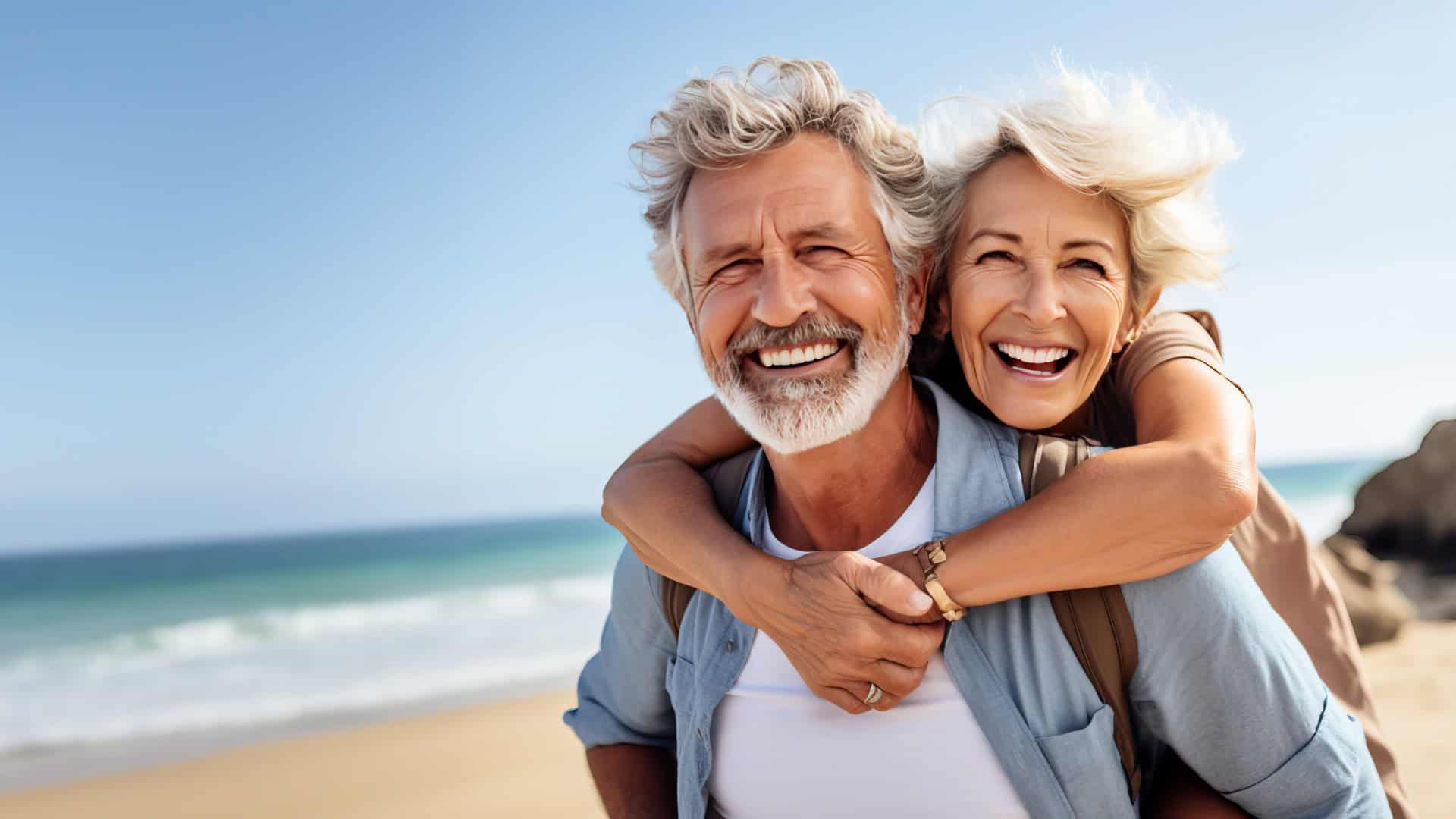 Älterer Mann und Frau am Strand, Mann trägt Frau Huckepack, beide lachen in die Kamera, Best Ager