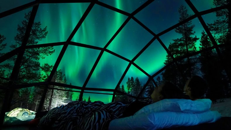 Ein Pärchen in einem Glasiglu in Lappland, das die grünen Polarlichter vom Bett aus bewundert