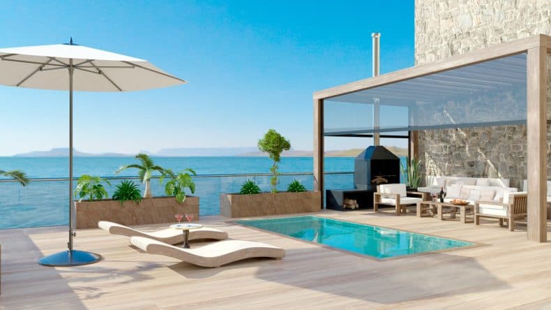 Blick auf eine hölzerne Terrasse mit Sonnenschirm, zwei modernen Liegen, Pool, einer hellen Sitzecke, blaues Meer im Hintergrund