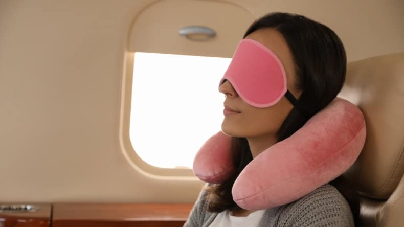 Frau sitzt entspannt mit Schlafmaske und Nackenkissen im Flugzeug